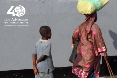 [ai] A woman carrying a large load on her head walks along a street, while a child follows closely behind with their hands clasped behind their back.